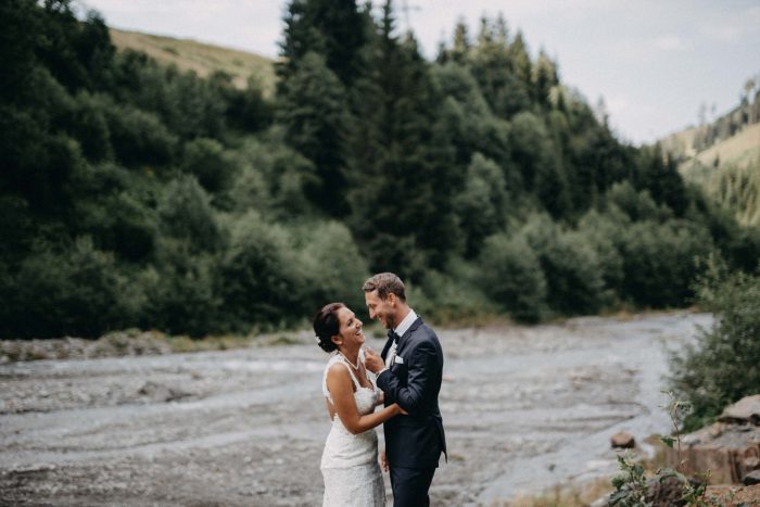 Brautpaar steht eng umarmt am Flussbett vor alpiner Bergkulisse – romantische Hochzeit in Saalbach-Hinterglemm inmitten der Natur