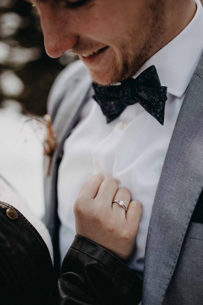 Detailaufnahme eines Bräutigams im Anzug mit Fliege, während die Braut seine Hand hält – romantische Winterhochzeit mit eleganten Hochzeitsdetails